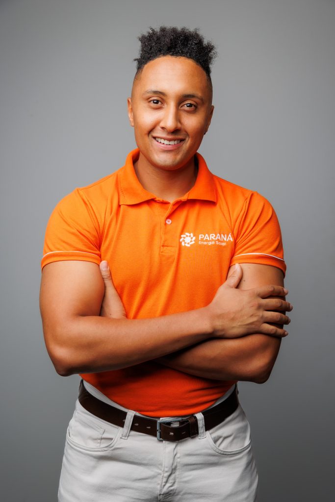 Homem com uniforme laranja sorrindo Alt text: Profissional da Paraná Energia Solar com uniforme laranja, sorrindo de braços cruzados, representando atendimento humanizado e confiança da empresa.